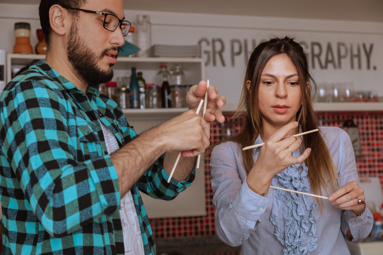 Un homme enseigne à une femme comment utiliser des baguettes dans une cuisine, symbolisant un moment d'apprentissage partagé.