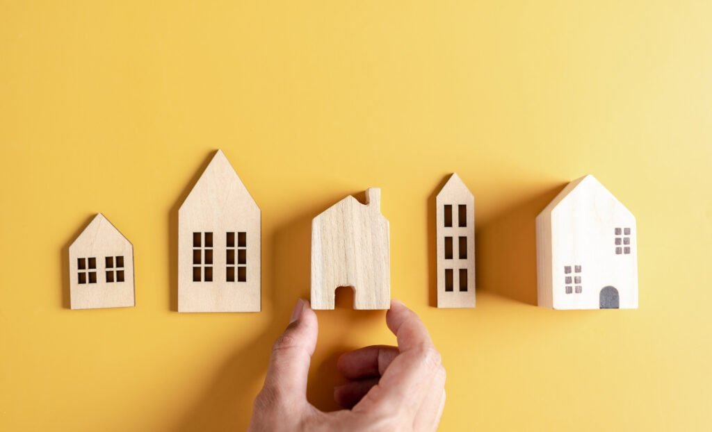 Une main tenant un modèle de maison en bois devant une sélection d'autres maisons en bois de formes différentes sur un fond jaune, symbolisant le processus de sélection de la maison parfaite.