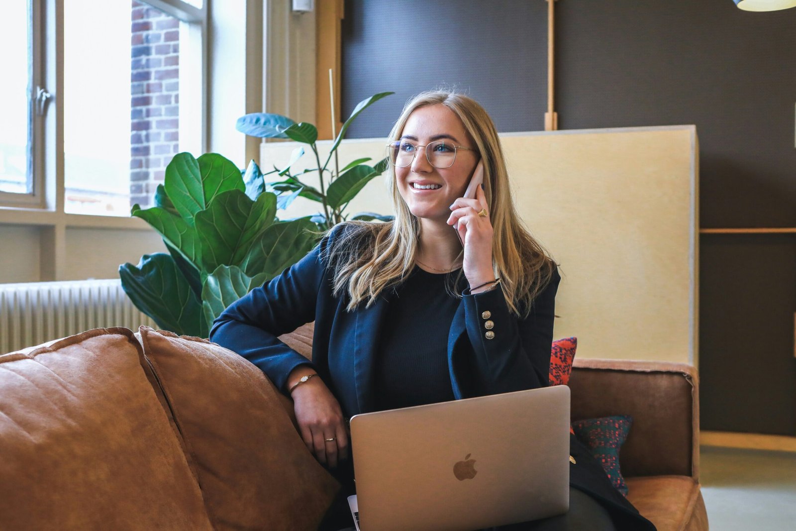 Femme d'affaires utilisant un téléphone et un ordinateur portable tout en souriant, assise dans un espace de bureau avec une plante verte en arrière-plan.
