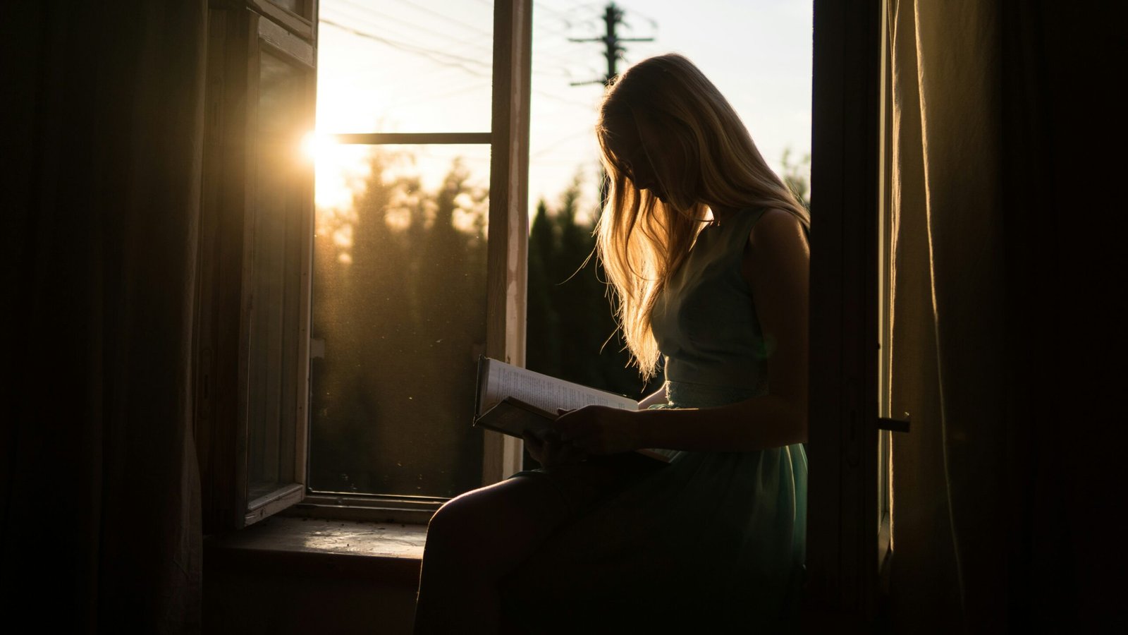 Contre-jour d'une femme lisant près d'une fenêtre au coucher du soleil, avec une lumière douce qui dessine sa silhouette.